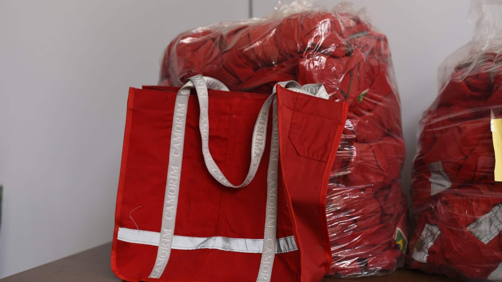 Ecobag vermelha com faixa refletiva e alças cinza escritas Camorim, feita de macacão de trabalho, na frente de sacos plásticos cheios de macacões vermelhos.