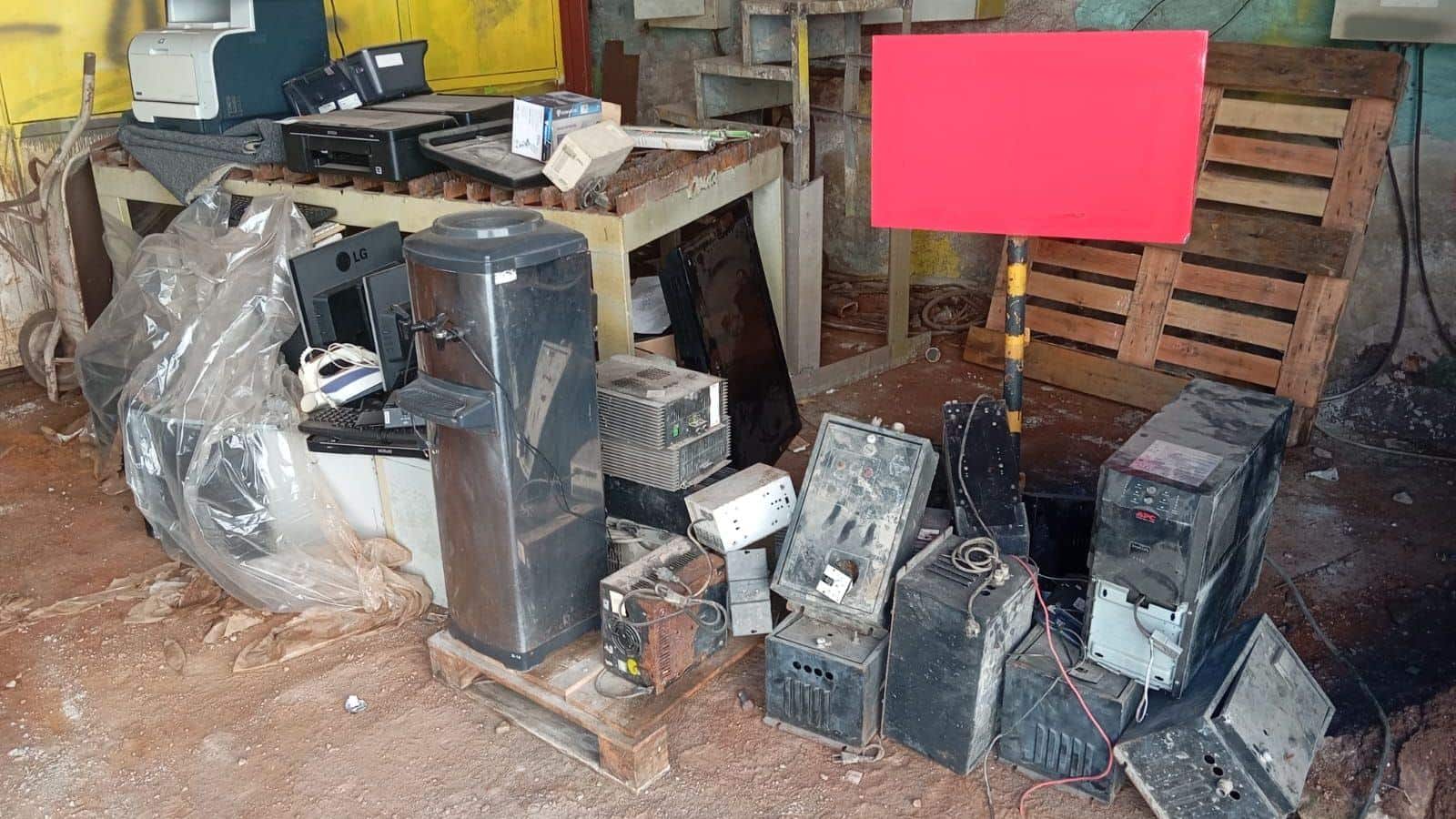 Aparelhos eletrônicos velhos amontoados no chão de um galpão: computadores, nobreaks, impressoras, um monitor e um bebedouro.