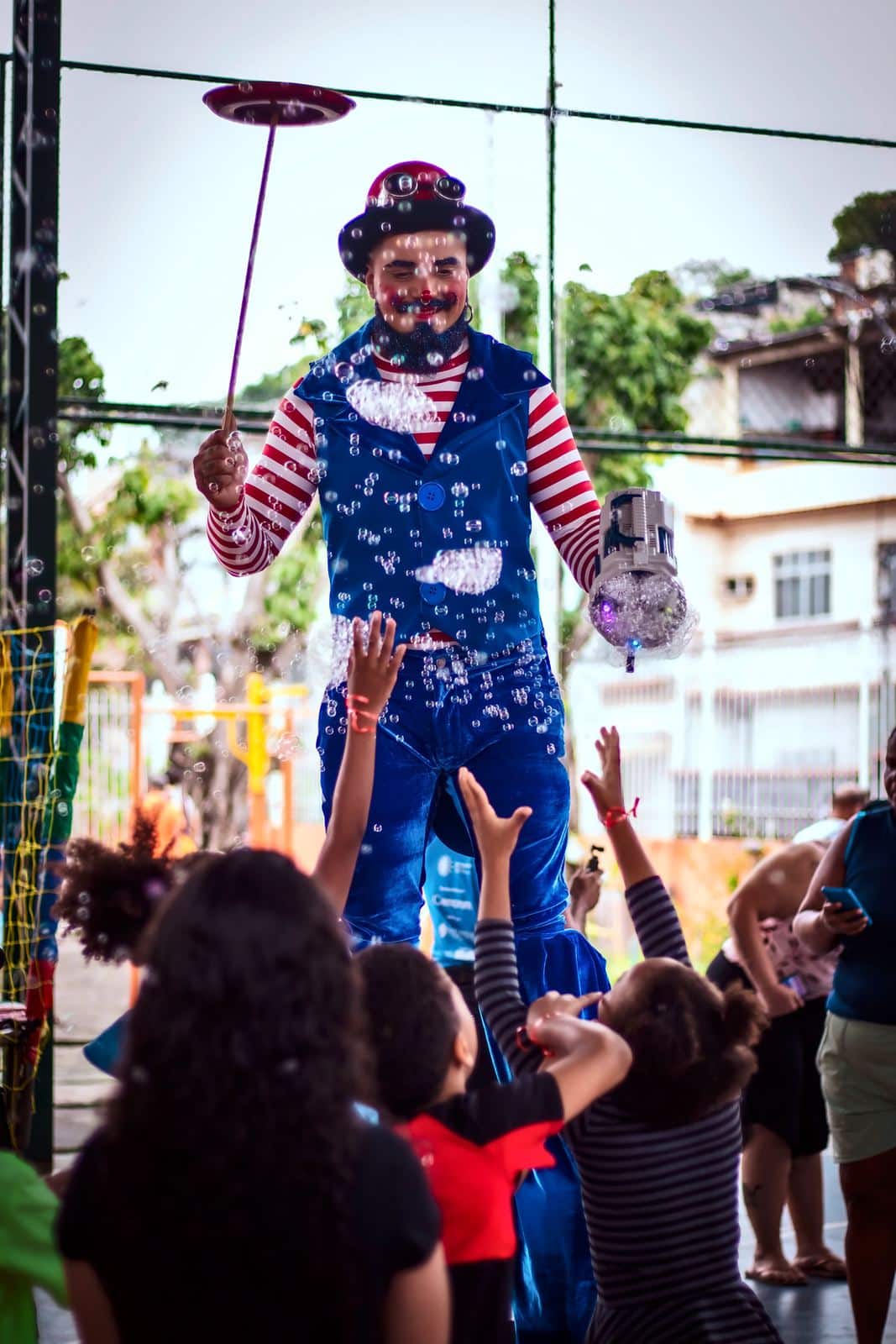 Artista de pernas de pau gira um prato no ar enquanto crianças estendem as mãos