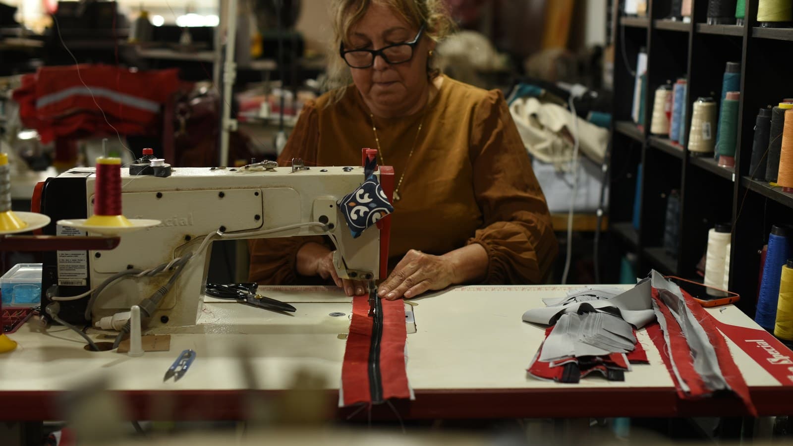 Costureira de óculos une tiras de tecido vermelho com zíper numa máquina de costura industrial; na mesa, retalhos vermelhos e cinza, e ao fundo uma estante de linhas coloridas.