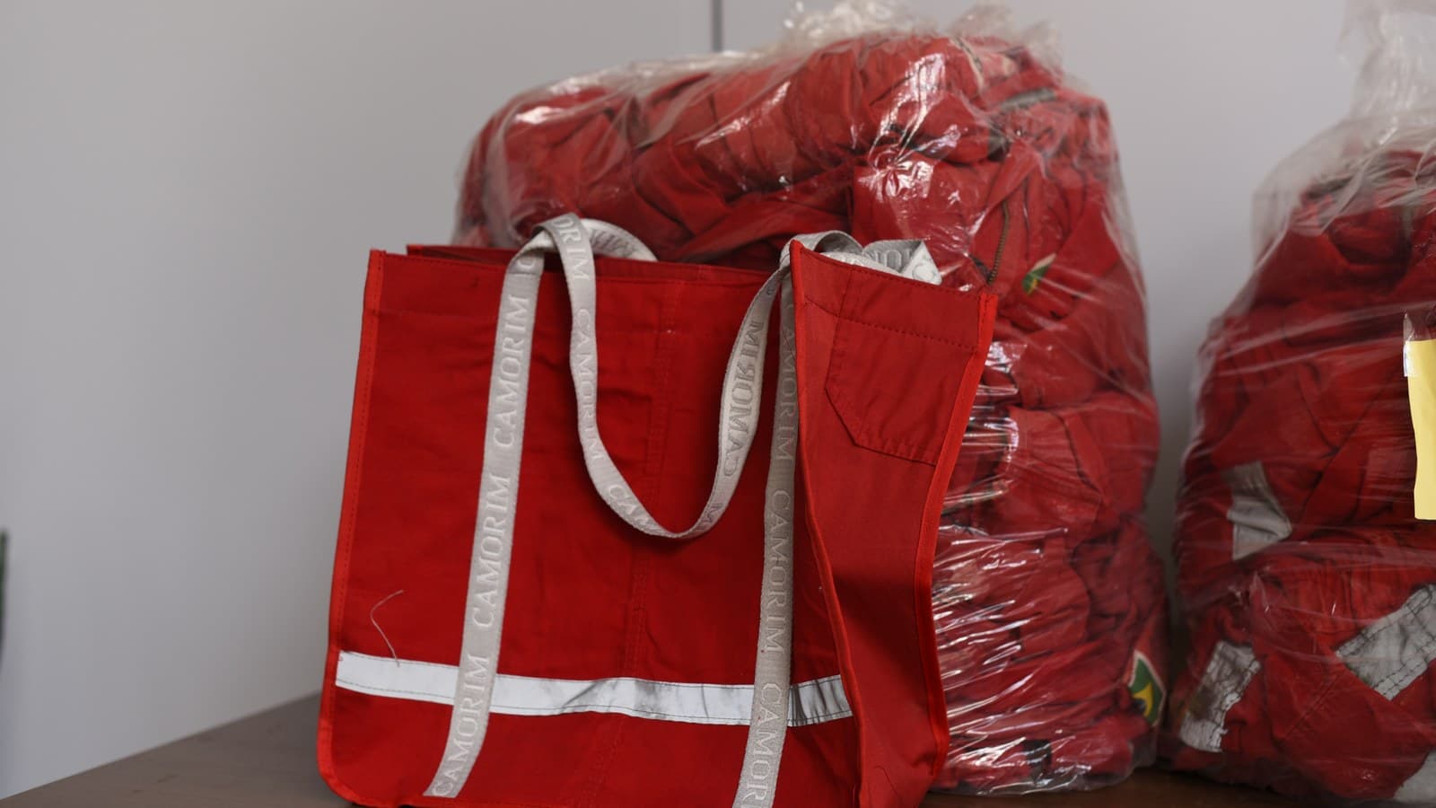 Ecobag vermelha com faixa refletiva e alças cinza escritas Camorim, feita de macacão de trabalho, na frente de sacos plásticos cheios de macacões vermelhos.