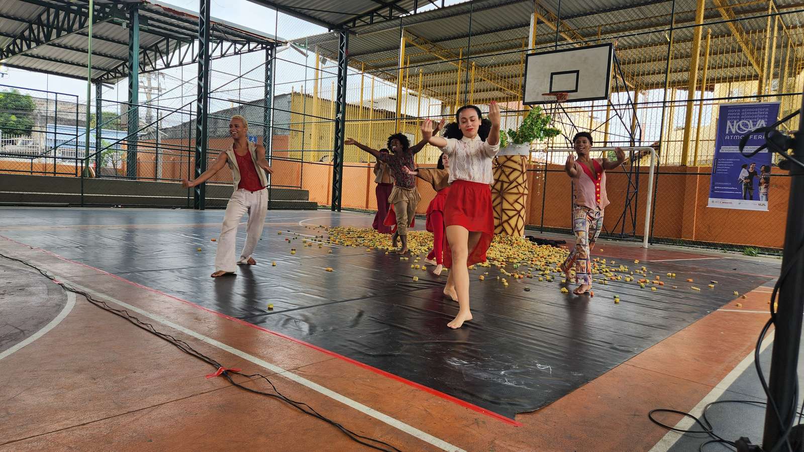Bailarinos dançam descalços sobre uma lona preta na quadra coberta de uma escola; ao fundo, um banner da Nova Atmosfera preso na grade.