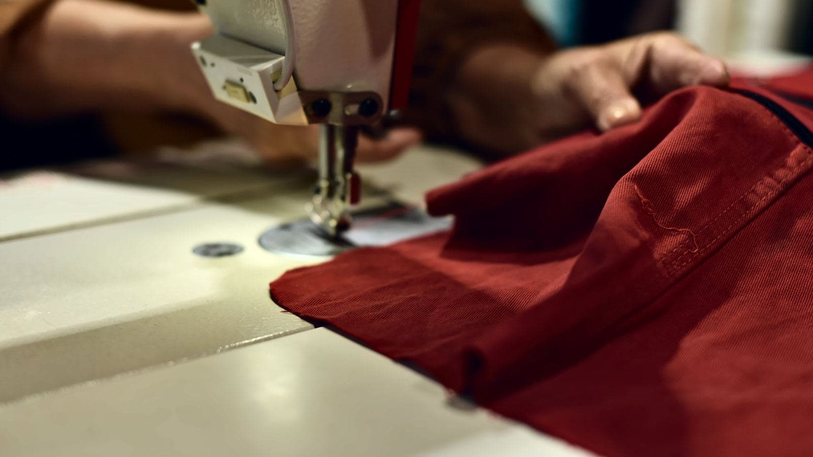 Mãos guiam um tecido vermelho sob a agulha de uma máquina de costura industrial.