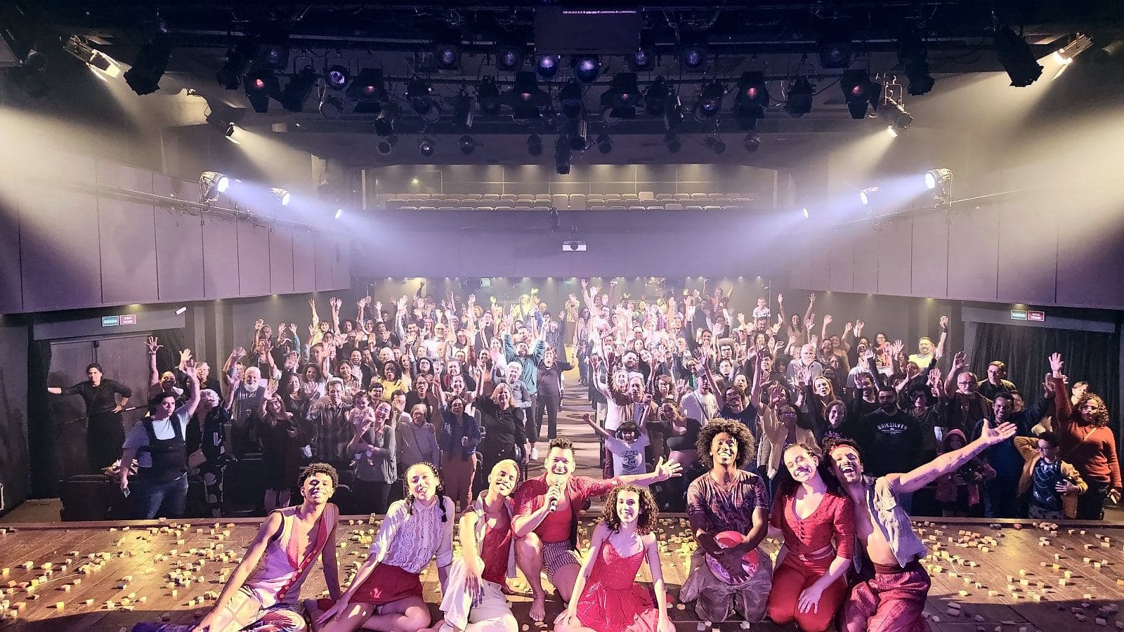 Elenco de bailarinos em figurino vermelho e bege sentado na beira do palco, de braços abertos, com a plateia lotada de braços erguidos atrás, sob luzes de teatro.