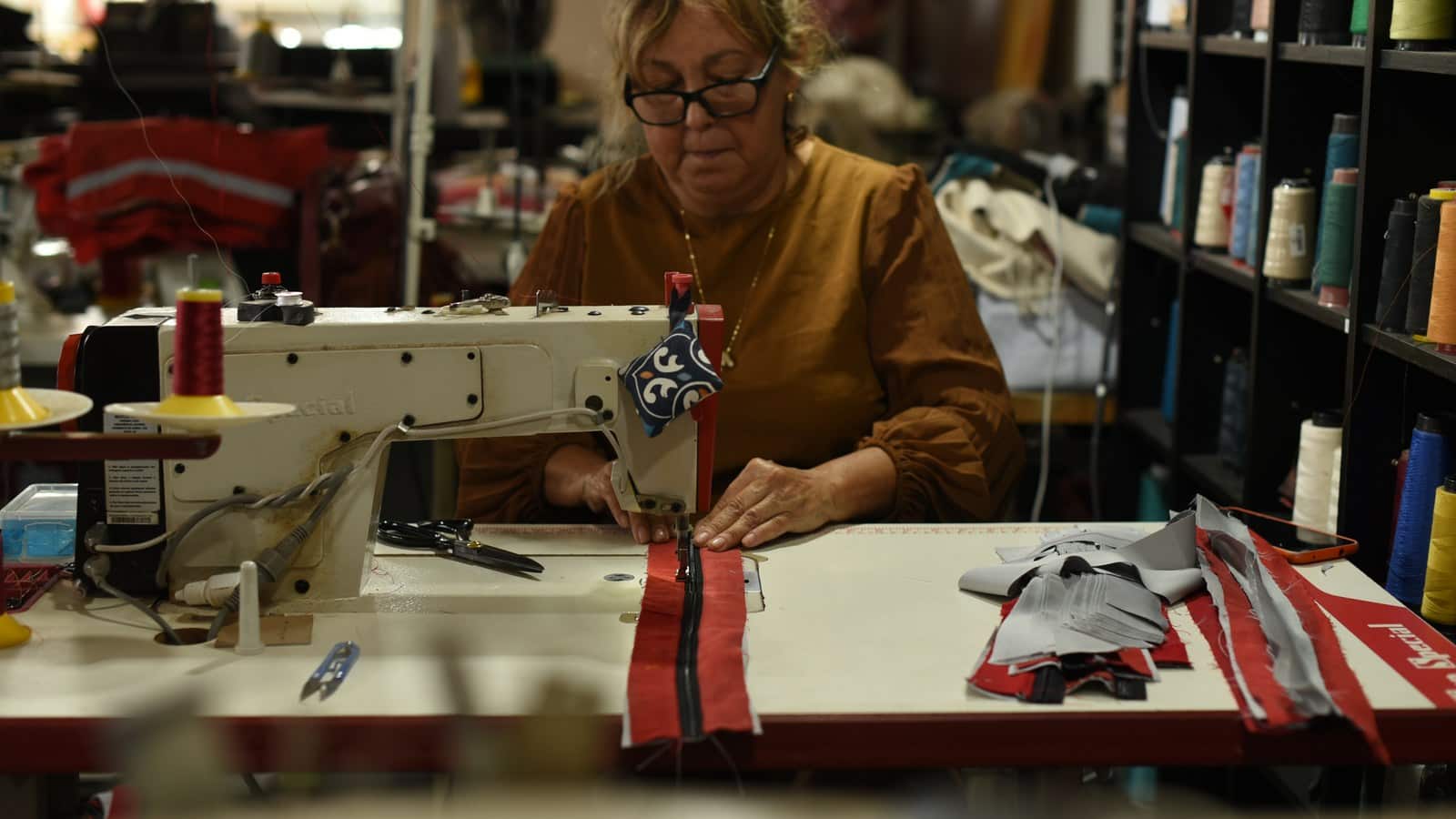Costureira de óculos une tiras de tecido vermelho com zíper numa máquina de costura industrial; na mesa, retalhos vermelhos e cinza, e ao fundo uma estante de linhas coloridas.