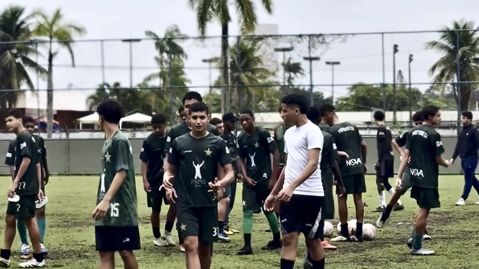 Jovens da base de uniforme verde com a marca Nova Atmosfera reunidos no campo, entre palmeiras, depois da rotina do grupo.