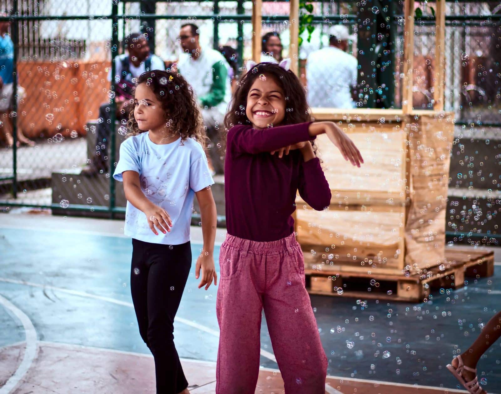 Duas meninas dançam e sorriem entre bolhas de sabão na quadra, no Dia das Crianças