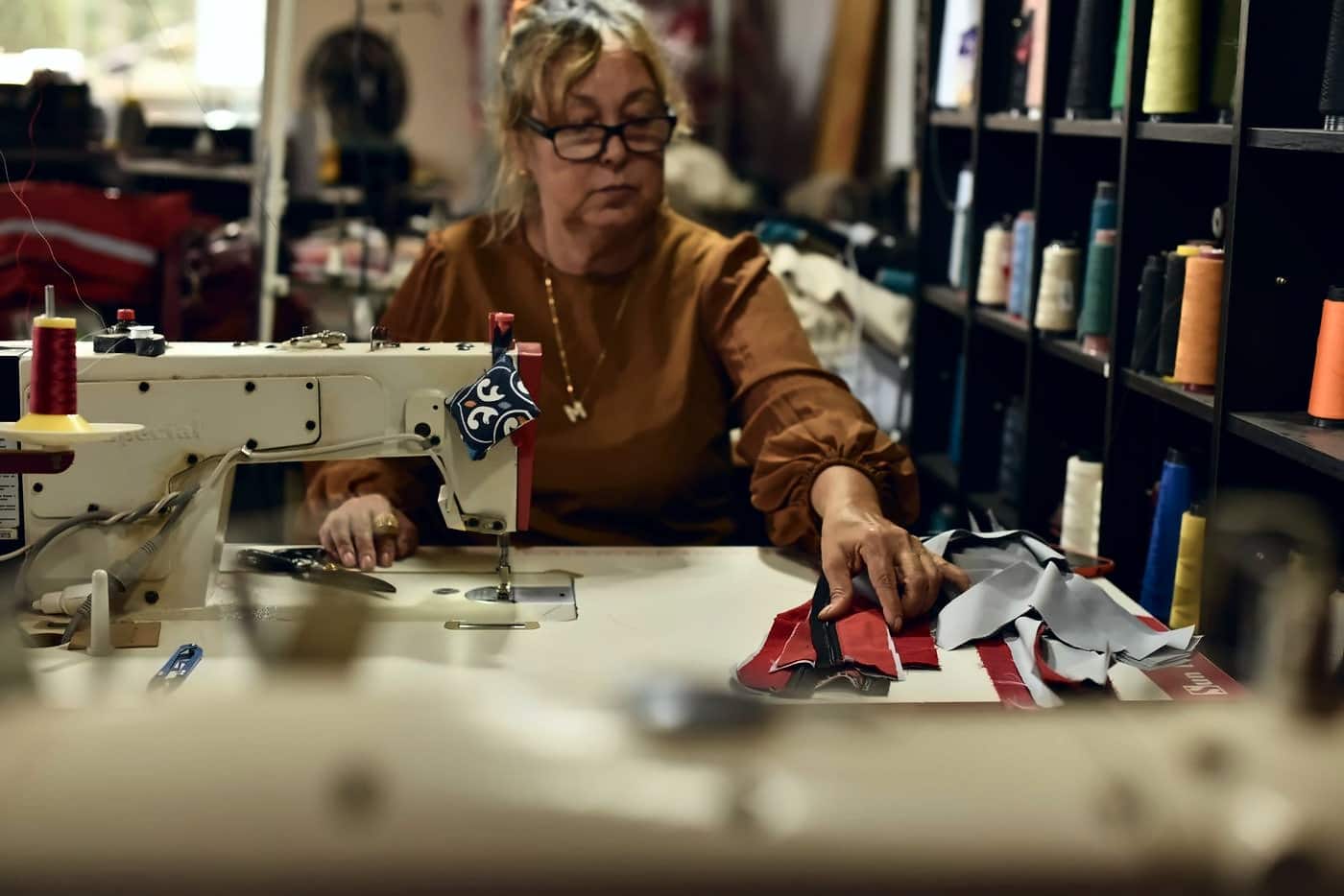 Costureira de óculos na máquina de costura, com retalhos vermelhos e cinza de macacões
